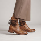 A pair of brown leather lace-up ankle boots worn with taupe trousers, shown from the calves down against a light gray studio background.