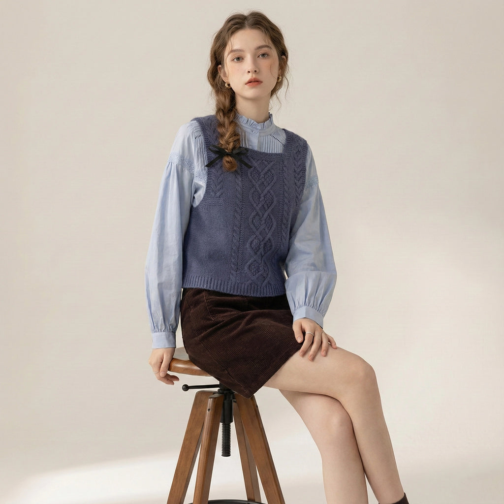 Woman sitting on a stool wearing a blue knitted vest over a light blue shirt and brown skirt.