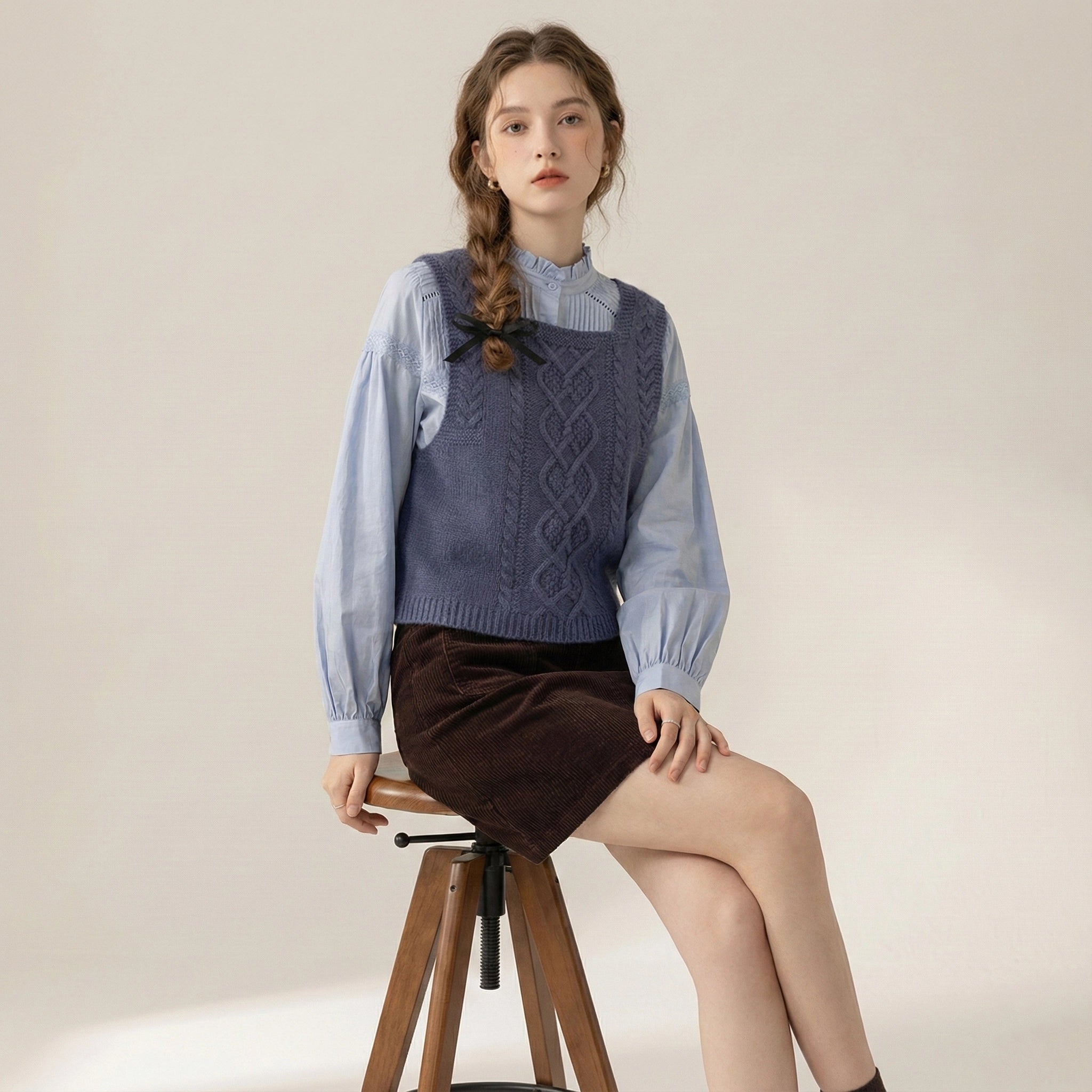 Woman sitting on a stool wearing a blue knitted vest over a light blue shirt and brown skirt.