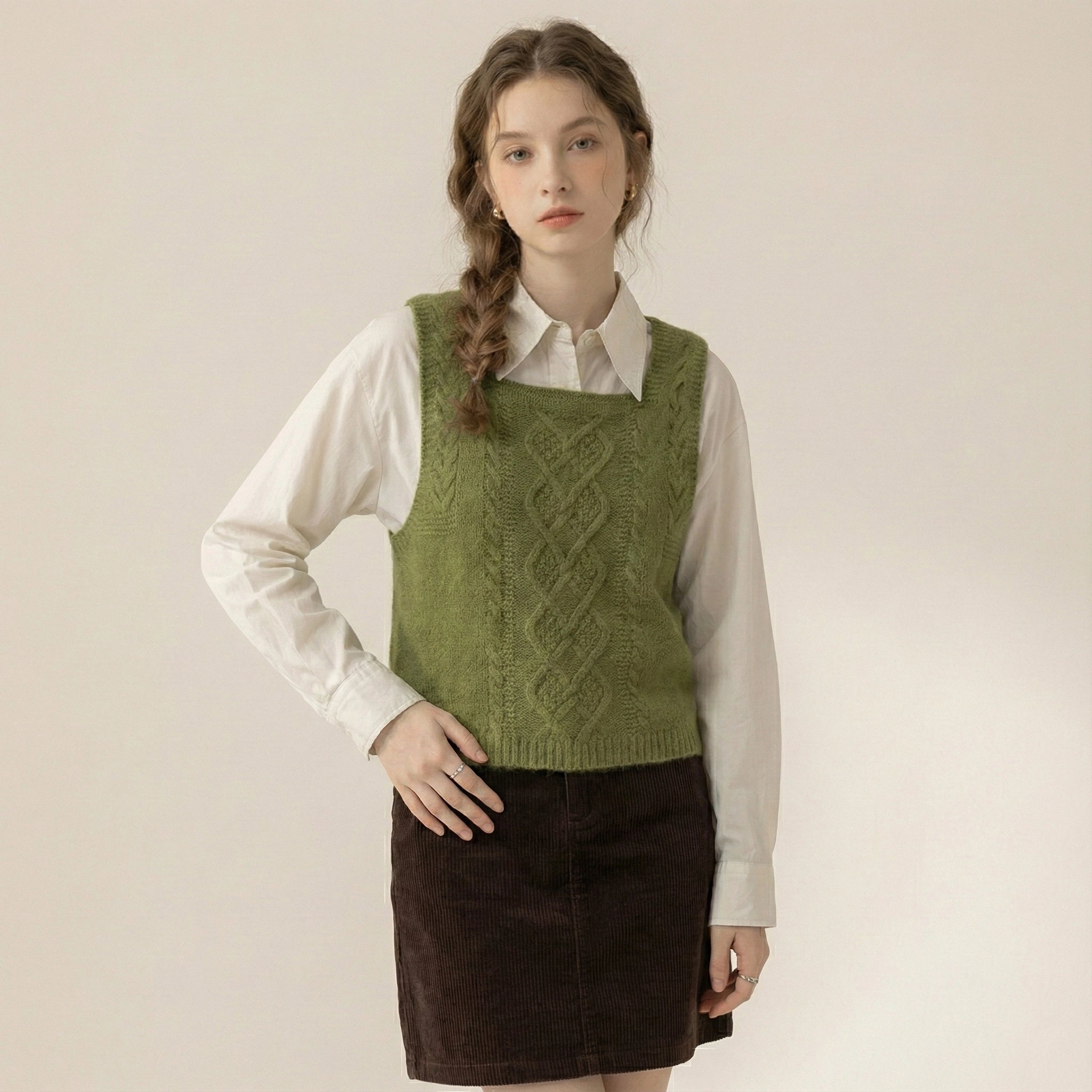 Woman wearing a green knitted vest over a white shirt and brown skirt on a beige background