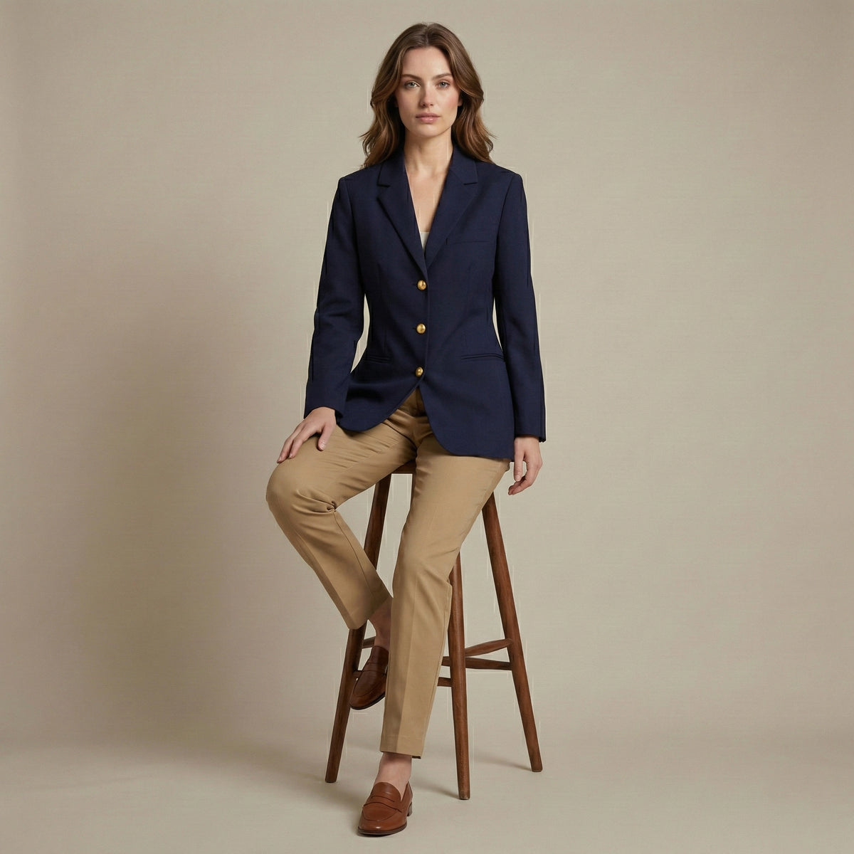 Woman wearing a navy blazer and beige pants sitting on a wooden stool against a beige background