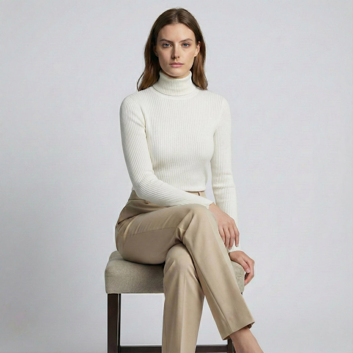 Woman wearing a white turtleneck sweater and beige pants sitting on a chair against a plain background