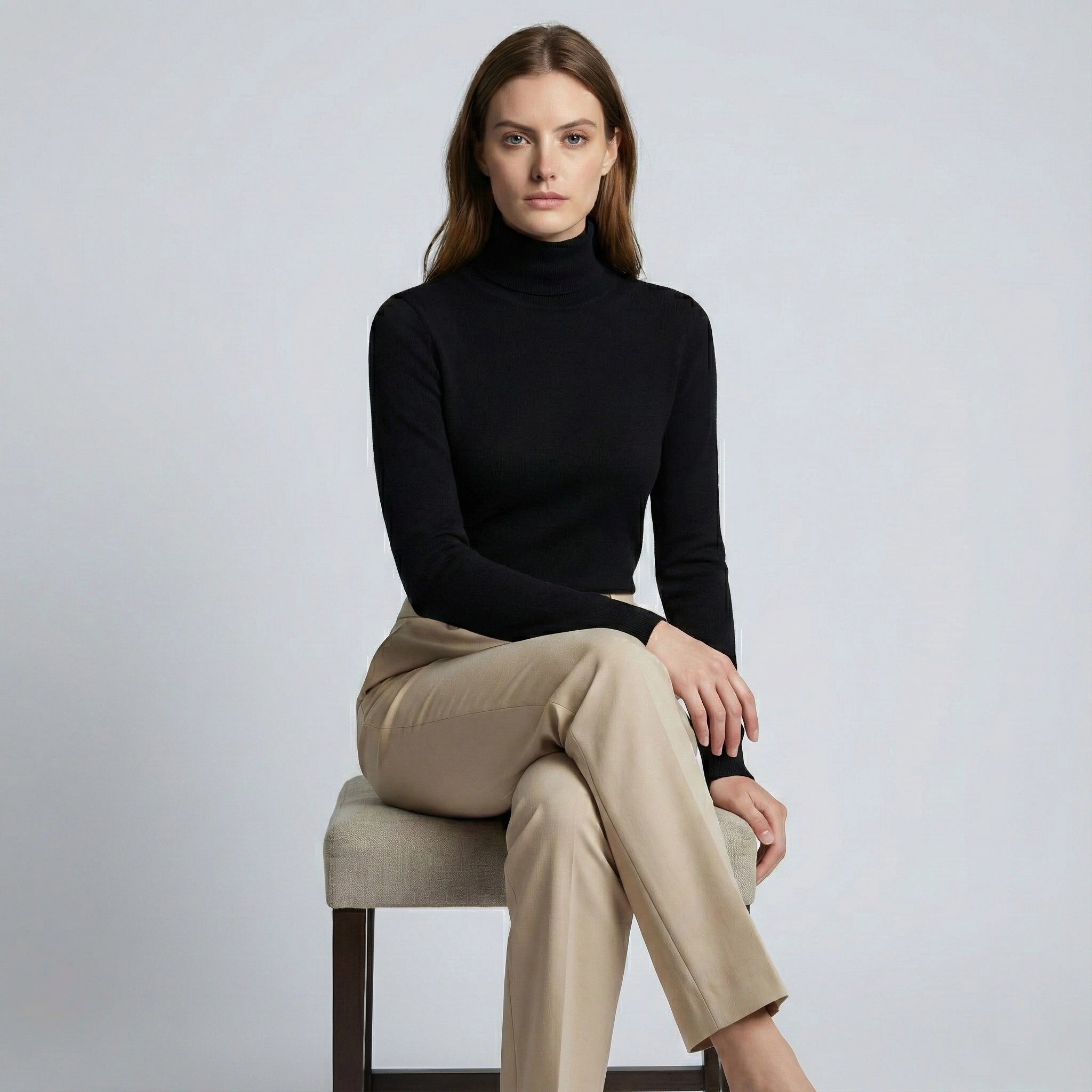 Woman wearing a black turtleneck and beige pants sitting on a chair against a plain background