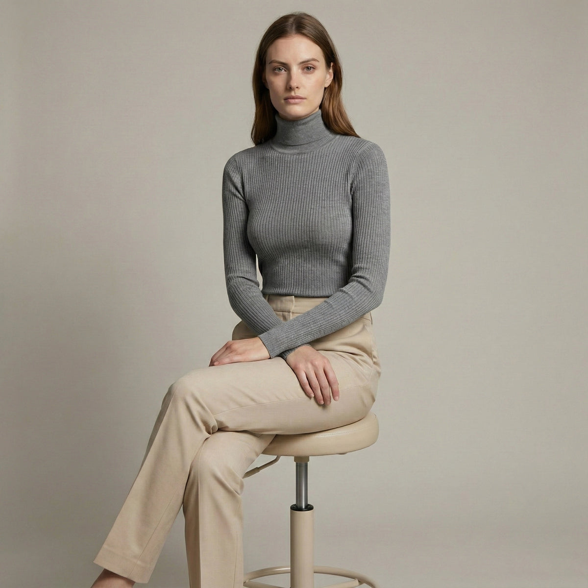 Woman sitting on a stool wearing a gray turtleneck and beige pants against a plain background