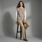 Woman sitting on a stool wearing a beige sweater vest, white shirt, tan pants, and brown shoes against a gray background.