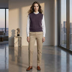 Woman wearing a purple sweater vest, white shirt, beige pants, and brown shoes in a modern room with large windows.