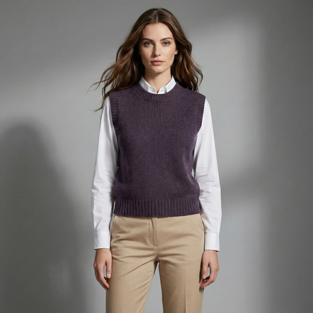 Woman wearing a purple sweater vest over a white shirt and beige pants on a gray background