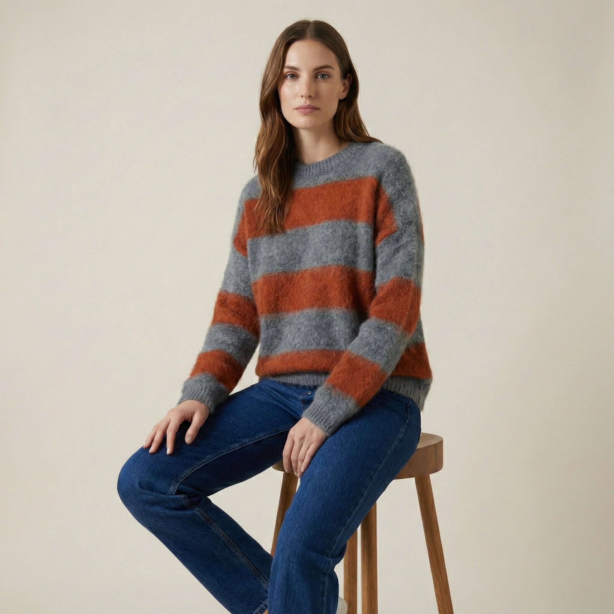 Woman wearing a striped sweater and jeans sitting on a stool against a plain background