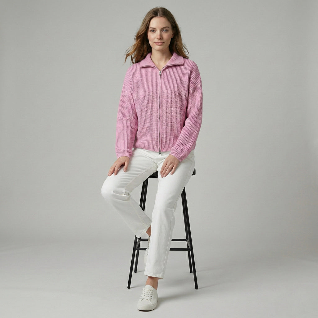 Woman wearing a pink jacket and white pants sitting on a black stool against a gray background