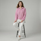 Woman wearing a pink jacket and white pants sitting on a black stool against a gray background