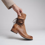 A hand wearing a beige sleeve holds up a light brown leather lace-up boot against a light gray studio background.