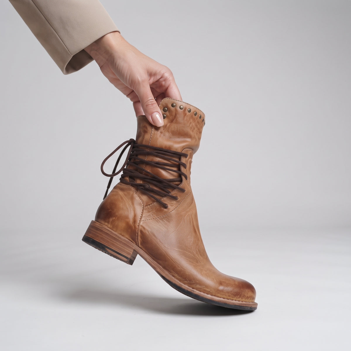 A hand wearing a beige sleeve holds up a light brown leather lace-up boot against a light gray studio background.