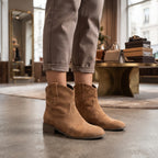 A person wearing tan suede ankle boots with a low block heel and pointed toes, paired with rolled-up taupe trousers in a stylish indoor boutique setting.