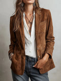 A woman wearing a brown suede blazer over a white button-down shirt and gray jeans, accessorized with layered necklaces and earrings, photographed against a light gray wall.