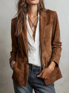 A woman wearing a brown suede blazer over a white button-down shirt and gray jeans, accessorized with layered necklaces and earrings, photographed against a light gray wall.