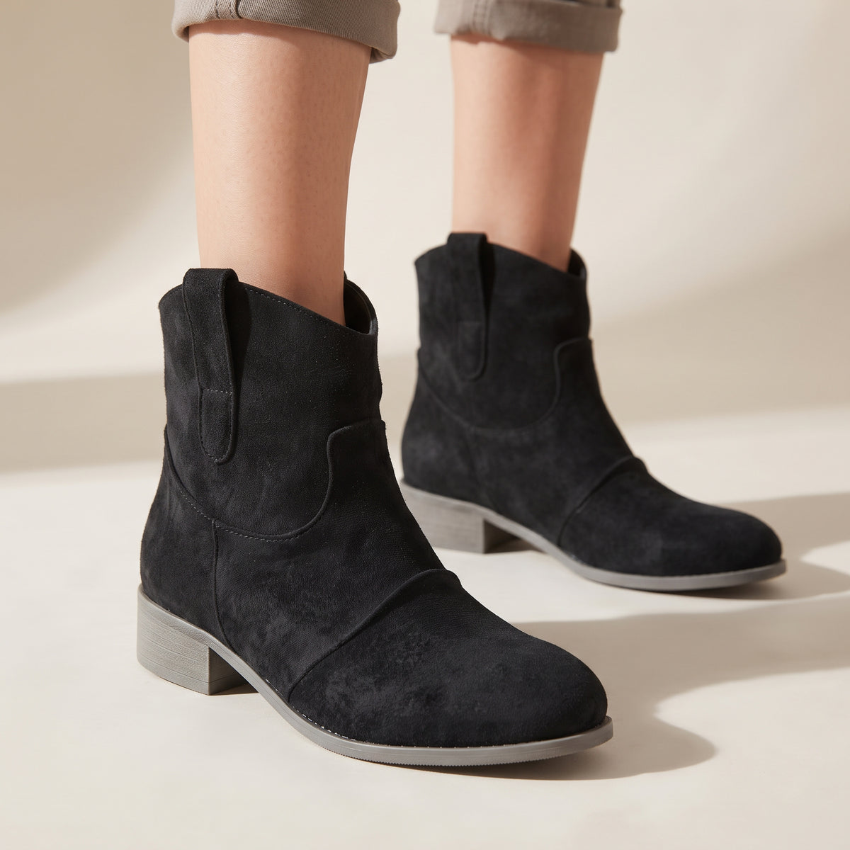 Black suede ankle boots with a low block heel, shown on a model wearing cuffed gray trousers against a warm beige background.