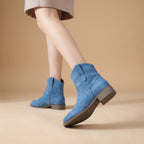 A person wearing light blue suede ankle boots with low brown block heels, shown from the knees down against a warm beige studio background.