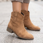 A person wearing tan suede ankle boots with a low block heel, shown from the calves down on a gray stone pavement.