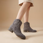 A person wearing gray suede ankle boots with low block heels, shown from the knees down against a warm beige studio background.
