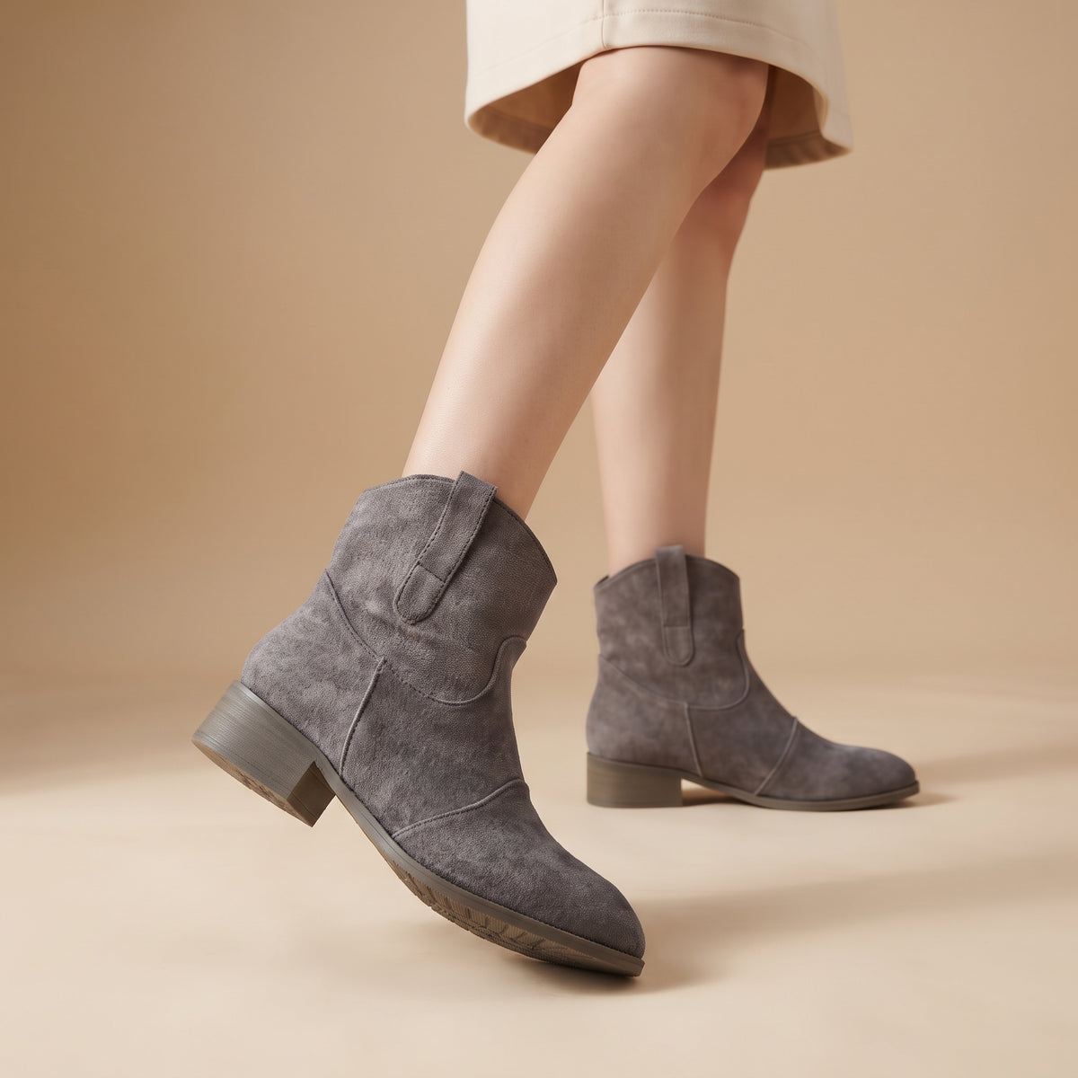 A person wearing gray suede ankle boots with low block heels, shown from the knees down against a warm beige studio background.