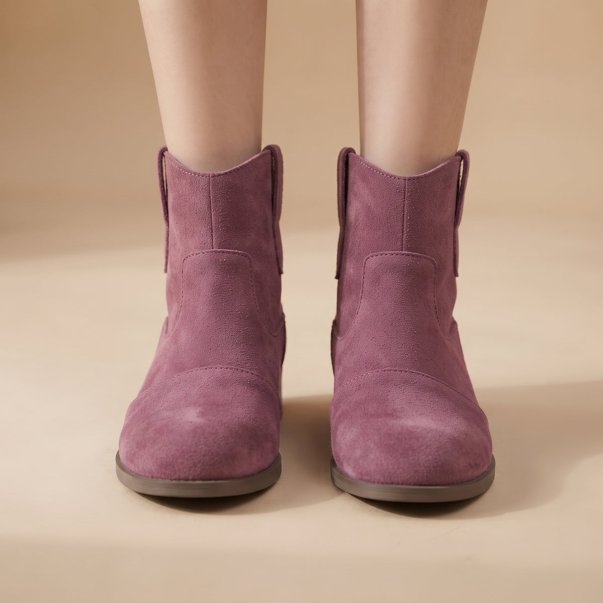 A pair of women's ankle boots in dusty mauve purple suede, shown on a model's feet against a warm beige background.