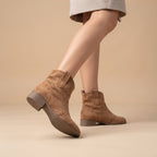 A person wearing taupe-brown suede ankle boots with elastic side panels and low block heels, shown against a plain beige studio background.