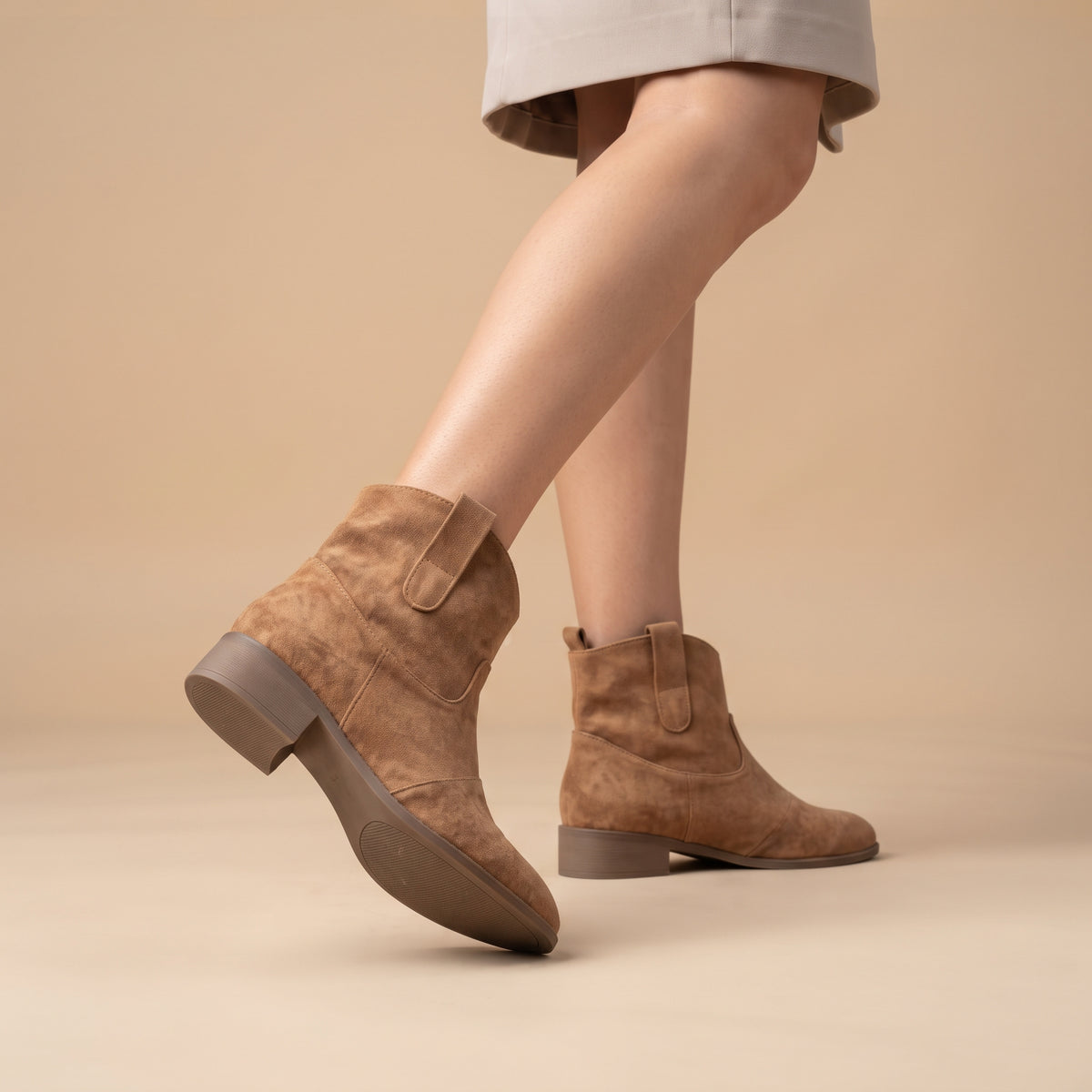 A person wearing taupe-brown suede ankle boots with elastic side panels and low block heels, shown against a plain beige studio background.