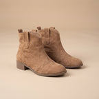 A pair of taupe suede ankle boots with elastic side panels and low block heels, displayed on a matching beige studio background.