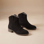 A pair of black suede ankle boots with low block heels, displayed on a warm beige studio background.