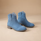 A pair of light blue suede ankle boots with low gray block heels, displayed on a warm beige studio background.