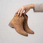 A pair of tan suede ankle boots held by a person's hand against a plain light gray background.