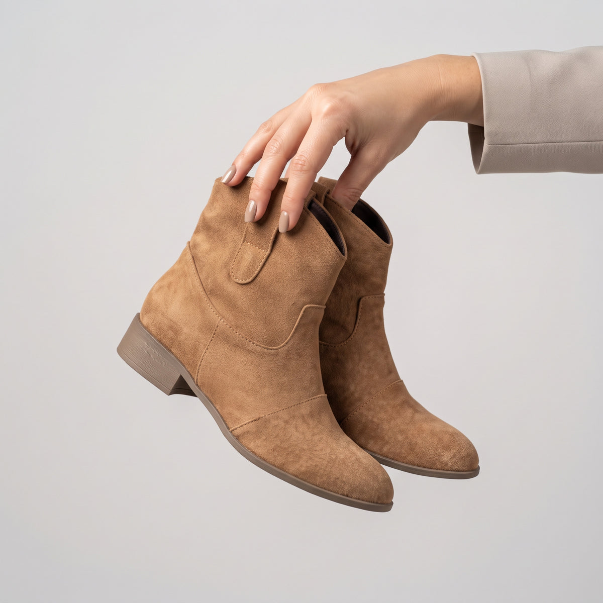 A pair of tan suede ankle boots held by a person's hand against a plain light gray background.