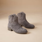 A pair of gray suede ankle boots with low block heels, shown on a warm beige studio background.