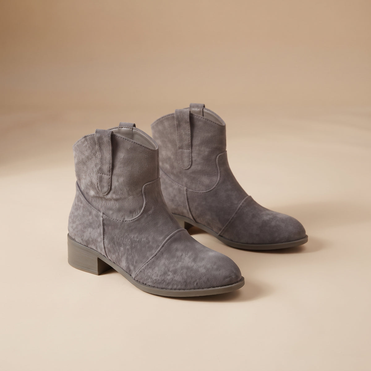 A pair of gray suede ankle boots with low block heels, shown on a warm beige studio background.