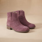 A pair of dusty purple suede ankle boots with low block heels, displayed on a warm beige background.