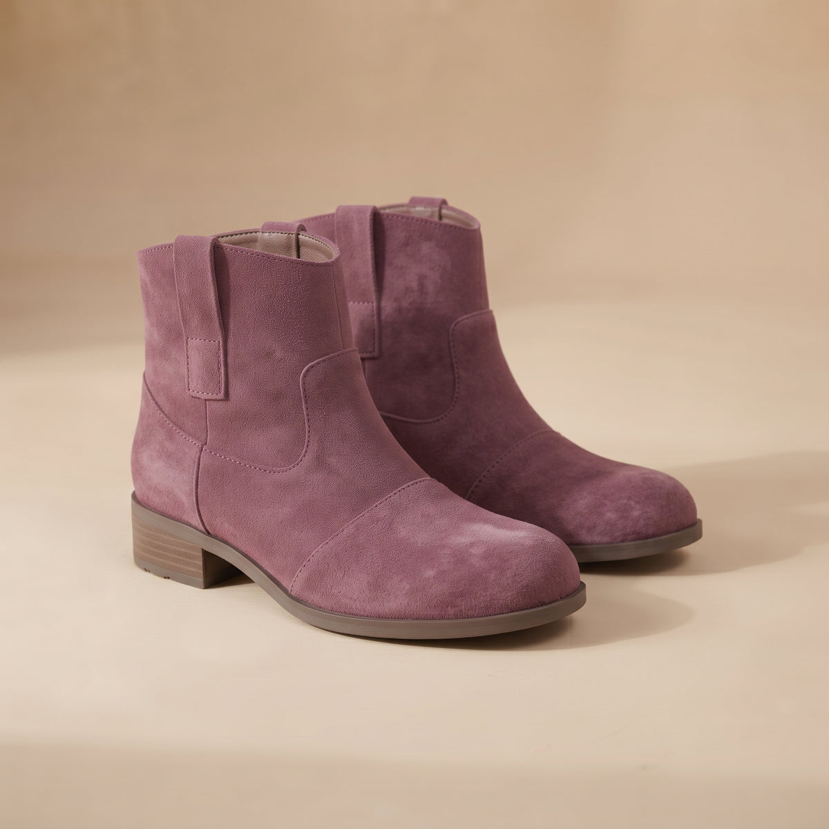 A pair of dusty purple suede ankle boots with low block heels, displayed on a warm beige background.